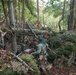 The image shows military personnel in full combat gear operating in a dense woodland environment. At least three soldiers are visible navigating through rocky, moss-covered terrain with fallen trees and leaf-covered ground. They appear to be conducting a field training exercise or tactical operation, carrying equipment and supplies.
