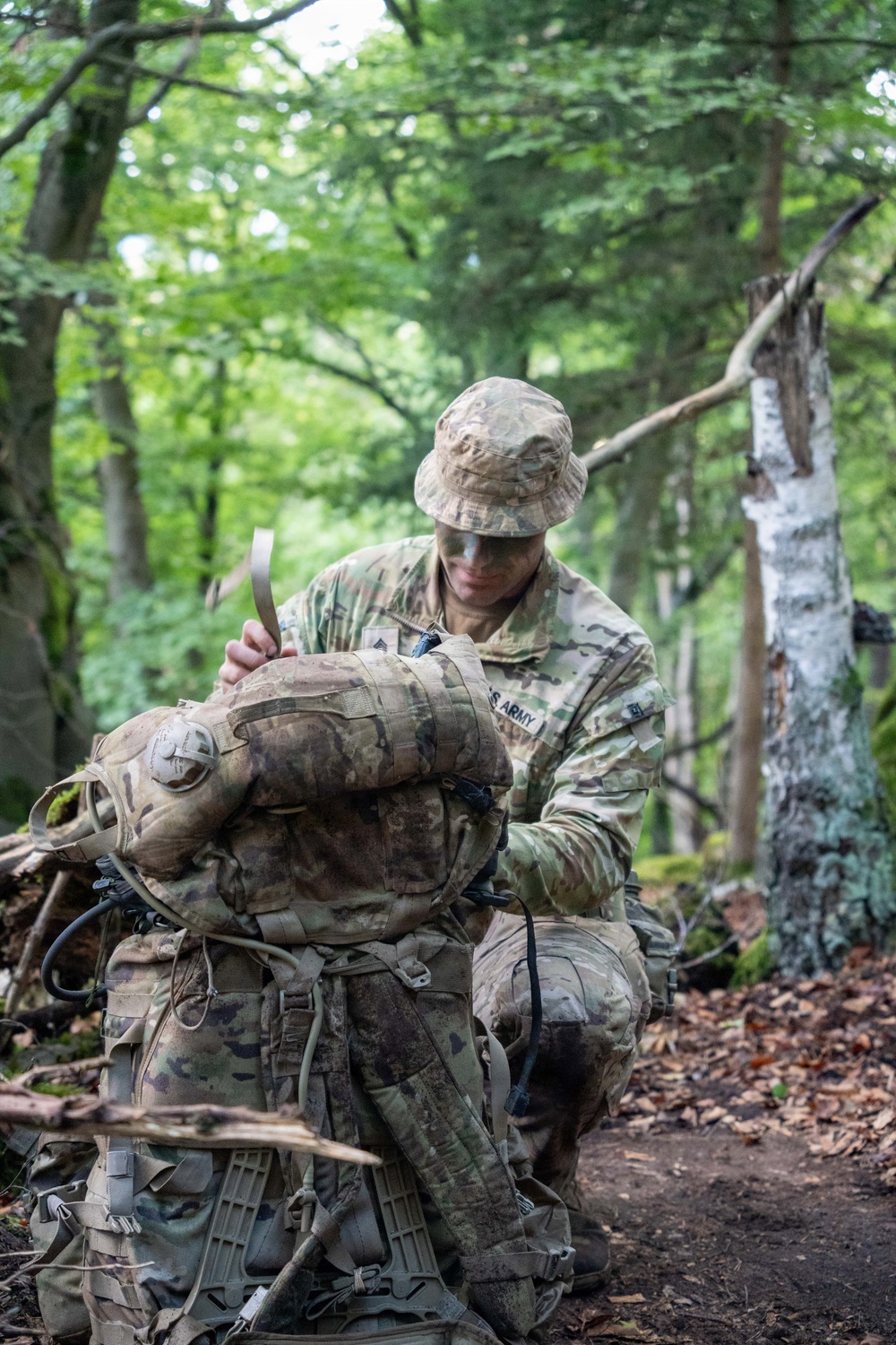 A U.S. Army soldier in Operational Camouflage Pattern (OCP) uniform and boonie hat kneels on a forest trail while adjusting or securing a large military rucksack. The soldier appears to be in a wooded training or operational environment with dense green foliage in the background. Face paint is visible on the soldier's face, suggesting a field training exercise or tactical operation.