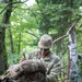 A U.S. Army soldier in Operational Camouflage Pattern (OCP) uniform and boonie hat kneels on a forest trail while adjusting or securing a large military rucksack. The soldier appears to be in a wooded training or operational environment with dense green foliage in the background. Face paint is visible on the soldier's face, suggesting a field training exercise or tactical operation.