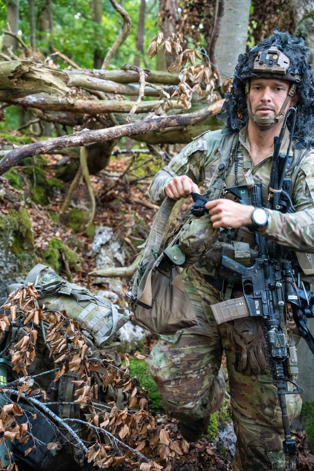 A soldier in full combat gear is positioned in a wooded forest environment, appearing to adjust or handle equipment straps. The individual is wearing MultiCam/OCP camouflage uniform with a tactical helmet equipped with a night vision mount and ghillie-style cover, along with a boom microphone headset. The setting suggests a military training exercise or field operation.