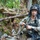 A soldier in full combat gear is positioned in a wooded forest environment, appearing to adjust or handle equipment straps. The individual is wearing MultiCam/OCP camouflage uniform with a tactical helmet equipped with a night vision mount and ghillie-style cover, along with a boom microphone headset. The setting suggests a military training exercise or field operation.