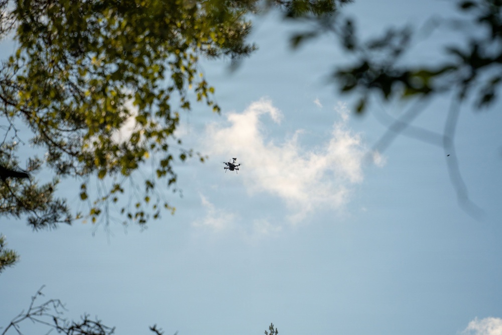 An upward-looking (worm's-eye view) photograph captures a small drone in flight against a partly cloudy blue sky. Tree branches with green foliage frame the upper-left and upper-right corners of the image. A large wind turbine is partially visible and out of focus on the right side of the frame.
