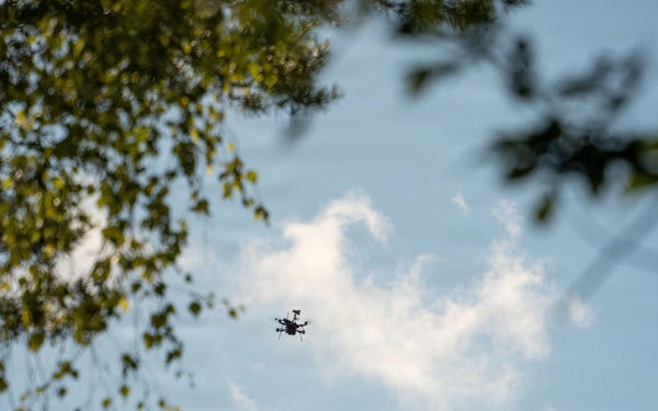 An upward-looking (worm's-eye view) photograph captures a small drone in flight against a partly cloudy blue sky. Tree branches with green foliage frame the upper-left and upper-right corners of the image. A large wind turbine is partially visible and out of focus on the right side of the frame.