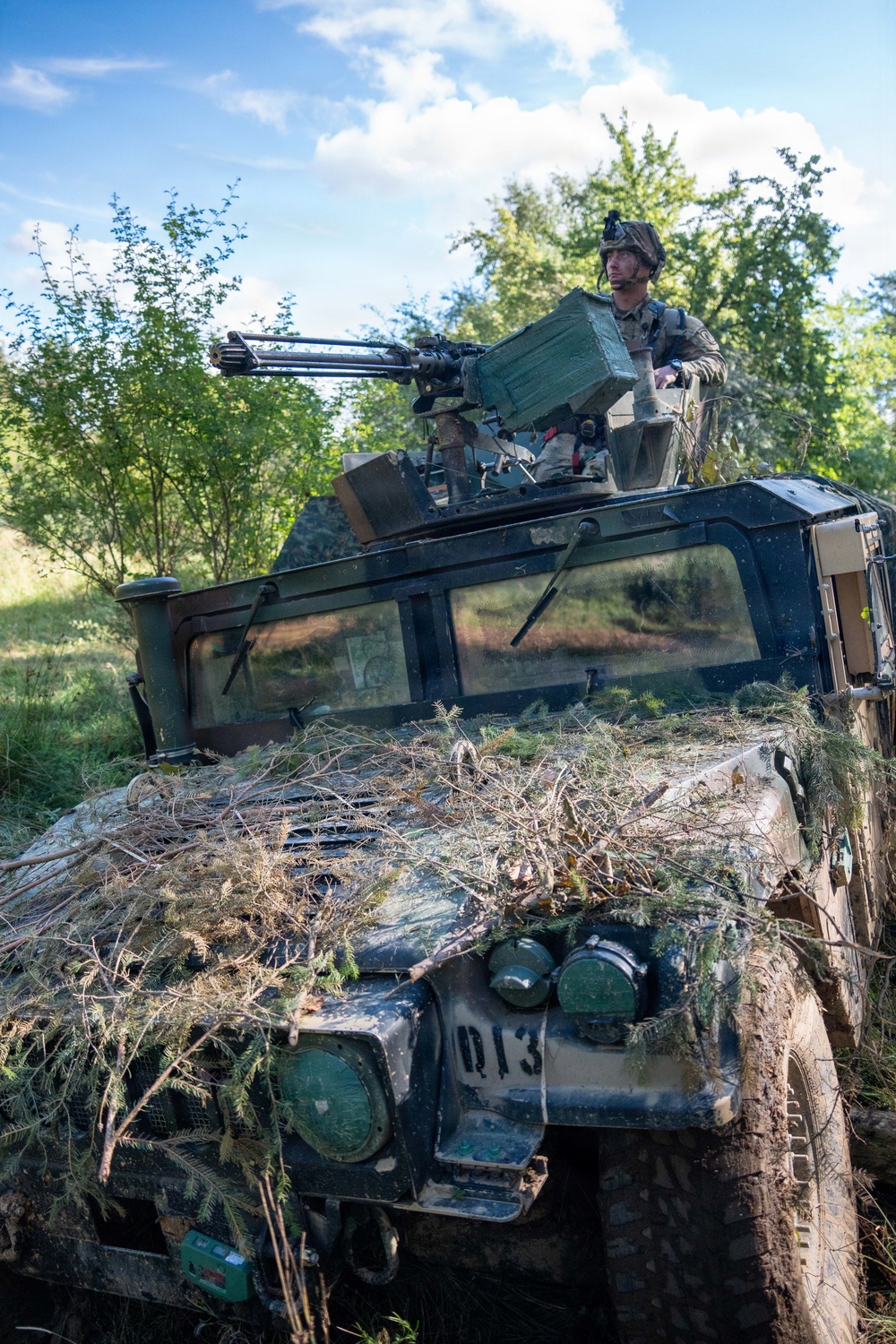 A military soldier in combat gear stands in the turret of a camouflaged HMMWV (Humvee) marked 'Q13', equipped with what appears to be an M134 Minigun or similar rotary weapon system. The vehicle is heavily adorned with natural vegetation (branches and foliage) for concealment purposes. The setting is a wooded, grassy area under a partly cloudy sky, suggesting a field training exercise or operational deployment.