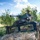 A military soldier in combat gear stands in the turret of a camouflaged HMMWV (Humvee) marked 'Q13', equipped with what appears to be an M134 Minigun or similar rotary weapon system. The vehicle is heavily adorned with natural vegetation (branches and foliage) for concealment purposes. The setting is a wooded, grassy area under a partly cloudy sky, suggesting a field training exercise or operational deployment.