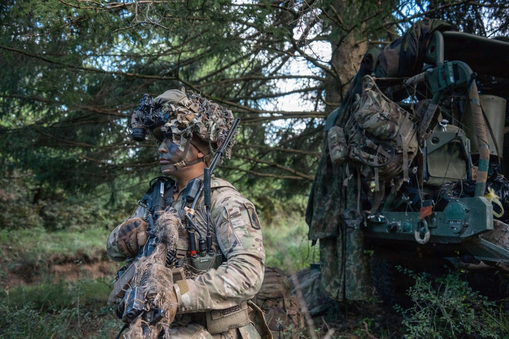 A heavily equipped soldier in full camouflage gear, including face paint and a ghillie-style helmet cover with night vision device mounted, stands alert in a wooded area. The soldier is holding a weapon and carrying a radio, positioned near what appears to be a military vehicle loaded with tactical gear and rucksacks. The setting suggests a field training exercise or operational deployment in a forested environment.