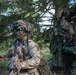 A heavily equipped soldier in full camouflage gear, including face paint and a ghillie-style helmet cover with night vision device mounted, stands alert in a wooded area. The soldier is holding a weapon and carrying a radio, positioned near what appears to be a military vehicle loaded with tactical gear and rucksacks. The setting suggests a field training exercise or operational deployment in a forested environment.