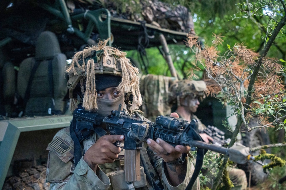Two heavily camouflaged soldiers in full tactical gear are positioned near a military vehicle in a wooded environment. The foreground soldier, wearing a US flag patch and ghillie-style helmet cover with face mask, is aiming an assault rifle. The scene appears to be a military training exercise or field operation.