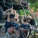 Two heavily camouflaged soldiers in full tactical gear are positioned near a military vehicle in a wooded environment. The foreground soldier, wearing a US flag patch and ghillie-style helmet cover with face mask, is aiming an assault rifle. The scene appears to be a military training exercise or field operation.