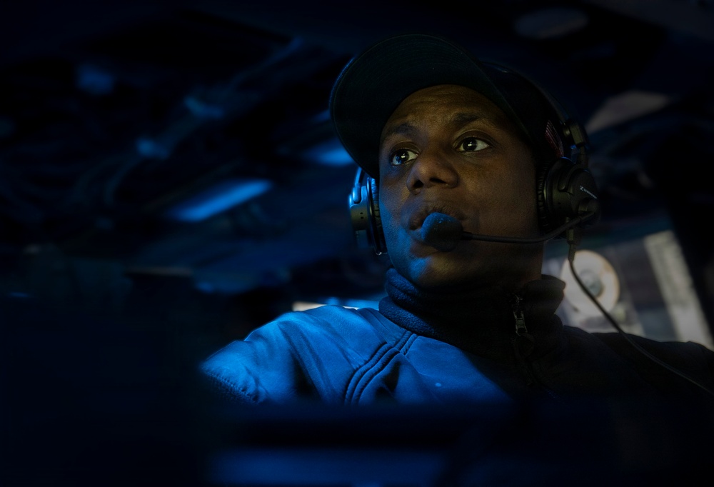 A person wearing a communications headset and dark baseball cap is illuminated by blue light in what appears to be a dimly lit operations or command environment. The individual appears to be actively monitoring or communicating, looking upward and to the side with focused attention. The dramatic blue lighting and dark surroundings suggest a military, naval, or aviation operations setting.