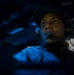 A person wearing a communications headset and dark baseball cap is illuminated by blue light in what appears to be a dimly lit operations or command environment. The individual appears to be actively monitoring or communicating, looking upward and to the side with focused attention. The dramatic blue lighting and dark surroundings suggest a military, naval, or aviation operations setting.