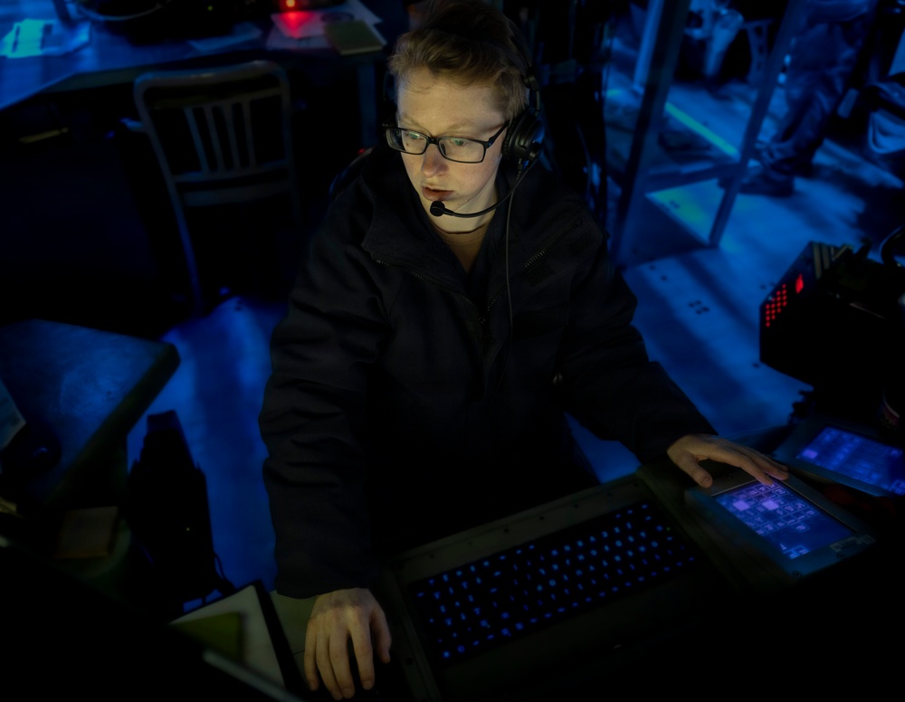 A young person wearing glasses and a headset with microphone is working at what appears to be a technical workstation in a darkened environment. The scene is illuminated primarily by blue ambient lighting and the glow of electronic displays. The setting suggests a live event production, gaming, or broadcast technical operations context.