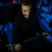 A young person wearing glasses and a headset with microphone is working at what appears to be a technical workstation in a darkened environment. The scene is illuminated primarily by blue ambient lighting and the glow of electronic displays. The setting suggests a live event production, gaming, or broadcast technical operations context.