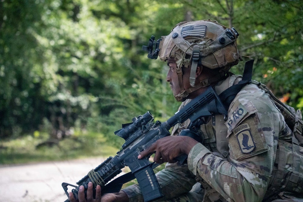 A U.S. Army soldier assigned to the 173rd Airborne Brigade is photographed in a wooded outdoor environment holding an assault rifle in a ready position. The soldier is wearing full combat gear including a multicam helmet with a U.S. flag patch and night vision mount, and an OCP (Operational Camouflage Pattern) uniform. The image appears to be taken during a training exercise or field operation.