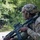 A U.S. Army soldier assigned to the 173rd Airborne Brigade is photographed in a wooded outdoor environment holding an assault rifle in a ready position. The soldier is wearing full combat gear including a multicam helmet with a U.S. flag patch and night vision mount, and an OCP (Operational Camouflage Pattern) uniform. The image appears to be taken during a training exercise or field operation.
