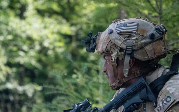 A U.S. Army soldier assigned to the 173rd Airborne Brigade is photographed in a wooded outdoor environment holding an assault rifle in a ready position. The soldier is wearing full combat gear including a multicam helmet with a U.S. flag patch and night vision mount, and an OCP (Operational Camouflage Pattern) uniform. The image appears to be taken during a training exercise or field operation.