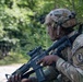 A U.S. Army soldier assigned to the 173rd Airborne Brigade is photographed in a wooded outdoor environment holding an assault rifle in a ready position. The soldier is wearing full combat gear including a multicam helmet with a U.S. flag patch and night vision mount, and an OCP (Operational Camouflage Pattern) uniform. The image appears to be taken during a training exercise or field operation.