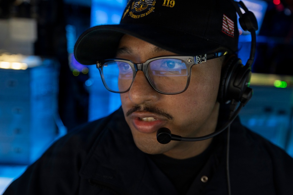 A young male military personnel wearing a black cap with the number '119' and an American flag patch is speaking into a communications headset with a boom microphone. He is wearing rectangular-framed glasses and a dark uniform, positioned in what appears to be a dimly lit operations or command center environment. The blue ambient lighting in the background suggests a naval or military command setting.