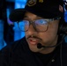 A young male military personnel wearing a black cap with the number '119' and an American flag patch is speaking into a communications headset with a boom microphone. He is wearing rectangular-framed glasses and a dark uniform, positioned in what appears to be a dimly lit operations or command center environment. The blue ambient lighting in the background suggests a naval or military command setting.