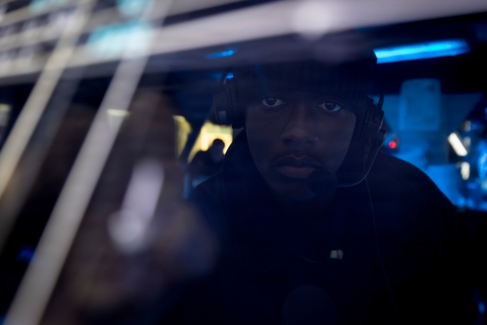 A person wearing a tactical headset or communications headgear is visible through a glass window or partition, illuminated by blue ambient lighting. The image is taken from outside looking in, with reflections visible on the glass surface. The environment behind the subject appears to be a dimly lit operational or technical space with blue lighting.