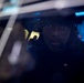 A person wearing a tactical headset or communications headgear is visible through a glass window or partition, illuminated by blue ambient lighting. The image is taken from outside looking in, with reflections visible on the glass surface. The environment behind the subject appears to be a dimly lit operational or technical space with blue lighting.
