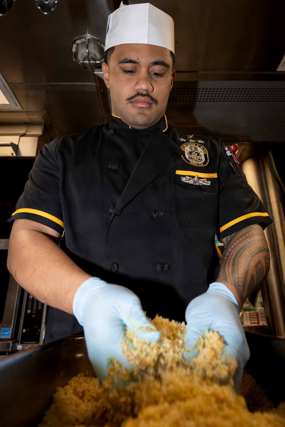 A U.S. Navy culinary specialist wearing a black chef's uniform with gold trim and a ship's patch (DDG-10 series) is preparing food in a shipboard galley. He is wearing blue latex gloves and mixing what appears to be rice or a grain-based dish in a large container. A traditional Polynesian-style sleeve tattoo is visible on his right arm.