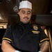 A U.S. Navy culinary specialist wearing a black chef's uniform with gold trim and a ship's patch (DDG-10 series) is preparing food in a shipboard galley. He is wearing blue latex gloves and mixing what appears to be rice or a grain-based dish in a large container. A traditional Polynesian-style sleeve tattoo is visible on his right arm.