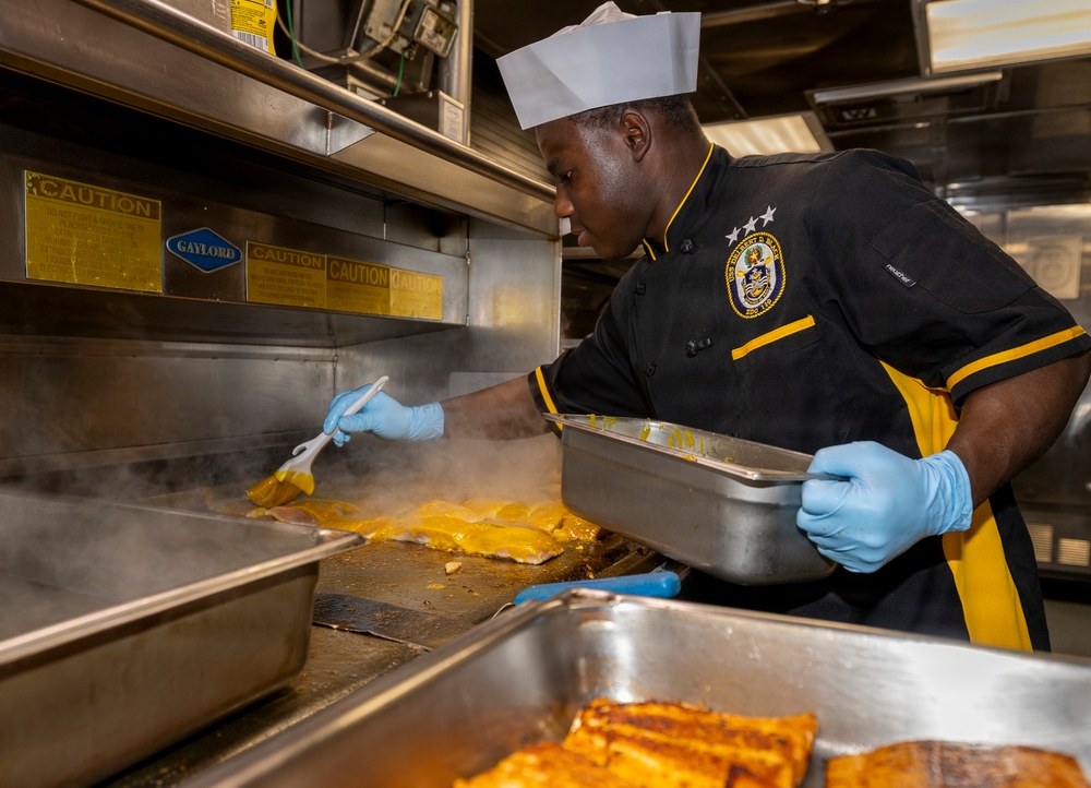 A chef in a black and gold uniform with a white toque is working in a professional kitchen, brushing a yellow sauce or glaze onto fish fillets on a flat-top grill. The cook is wearing blue nitrile gloves and holding a metal pan, with steam rising from the cooking surface. The setting appears to be a military galley kitchen, indicated by the uniform patch and 'Gaylord' ventilation equipment.