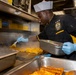 A chef in a black and gold uniform with a white toque is working in a professional kitchen, brushing a yellow sauce or glaze onto fish fillets on a flat-top grill. The cook is wearing blue nitrile gloves and holding a metal pan, with steam rising from the cooking surface. The setting appears to be a military galley kitchen, indicated by the uniform patch and 'Gaylord' ventilation equipment.