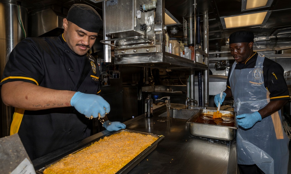 Two culinary workers in matching black and gold uniforms are preparing food in a professional kitchen environment, likely a military galley based on the insignia visible on their uniforms. The worker on the left is spreading or portioning a shredded/grated food item on a large baking sheet, while the worker on the right is basting or glazing a food item in a hotel pan. Both workers are wearing blue nitrile gloves and black skull caps.