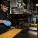 Two culinary workers in matching black and gold uniforms are preparing food in a professional kitchen environment, likely a military galley based on the insignia visible on their uniforms. The worker on the left is spreading or portioning a shredded/grated food item on a large baking sheet, while the worker on the right is basting or glazing a food item in a hotel pan. Both workers are wearing blue nitrile gloves and black skull caps.