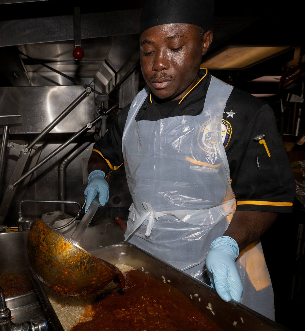 A military cook, wearing a black uniform with a ship's insignia patch, black chef's hat, blue latex gloves, and a clear plastic apron, is preparing food in what appears to be a naval vessel galley. The individual is actively stirring or transferring a large quantity of rice and tomato-based sauce from a large cooking vessel into a stainless steel pan. The environment is a professional industrial kitchen with stainless steel equipment visible in the background.