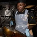 A military cook, wearing a black uniform with a ship's insignia patch, black chef's hat, blue latex gloves, and a clear plastic apron, is preparing food in what appears to be a naval vessel galley. The individual is actively stirring or transferring a large quantity of rice and tomato-based sauce from a large cooking vessel into a stainless steel pan. The environment is a professional industrial kitchen with stainless steel equipment visible in the background.