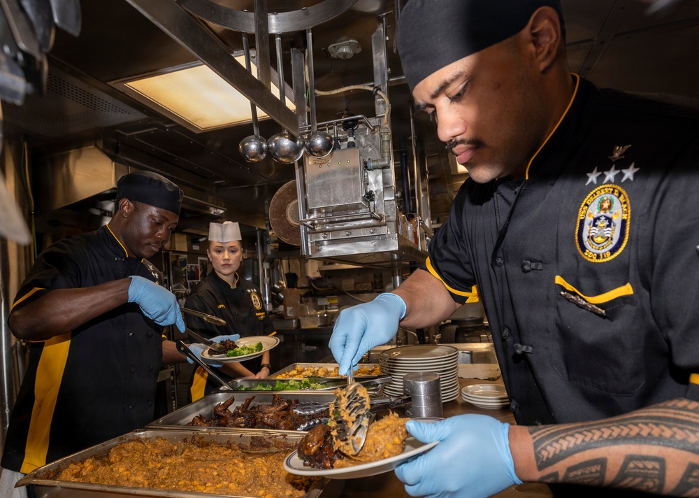 Three culinary specialists in matching black and gold uniforms are plating meals in a military ship's galley kitchen. The personnel are wearing blue nitrile gloves and working with trays of food including rice, braised meat, and vegetables. A ship's crest patch is visible on the uniform of the foreground individual, indicating this is a U.S. Navy vessel.