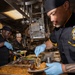 Three culinary specialists in matching black and gold uniforms are plating meals in a military ship's galley kitchen. The personnel are wearing blue nitrile gloves and working with trays of food including rice, braised meat, and vegetables. A ship's crest patch is visible on the uniform of the foreground individual, indicating this is a U.S. Navy vessel.