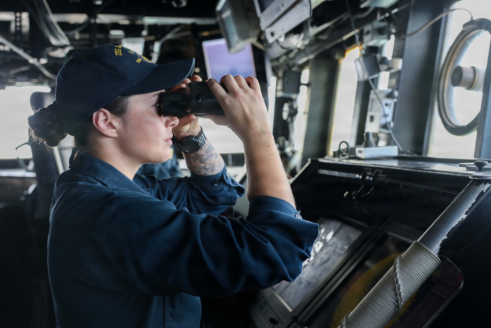 A naval service member in dark blue utility uniform is standing on the bridge of a warship, looking through binoculars. The individual is wearing a navy ball cap with gold lettering, suggesting a specific ship assignment. The bridge environment includes navigation equipment, monitors, and control panels typical of a U.S. Navy vessel.