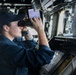 A naval service member in dark blue utility uniform is standing on the bridge of a warship, looking through binoculars. The individual is wearing a navy ball cap with gold lettering, suggesting a specific ship assignment. The bridge environment includes navigation equipment, monitors, and control panels typical of a U.S. Navy vessel.