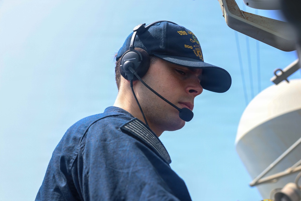 A young male U.S. Navy sailor is photographed on the deck of a naval vessel, wearing a dark blue uniform and a USS DDG ship's ball cap with gold lettering. He is equipped with a Sennheiser communications headset with a boom microphone, suggesting he is engaged in active communications duty. The background shows a clear blue sky and partial view of shipboard equipment.