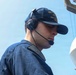 A young male U.S. Navy sailor is photographed on the deck of a naval vessel, wearing a dark blue uniform and a USS DDG ship's ball cap with gold lettering. He is equipped with a Sennheiser communications headset with a boom microphone, suggesting he is engaged in active communications duty. The background shows a clear blue sky and partial view of shipboard equipment.