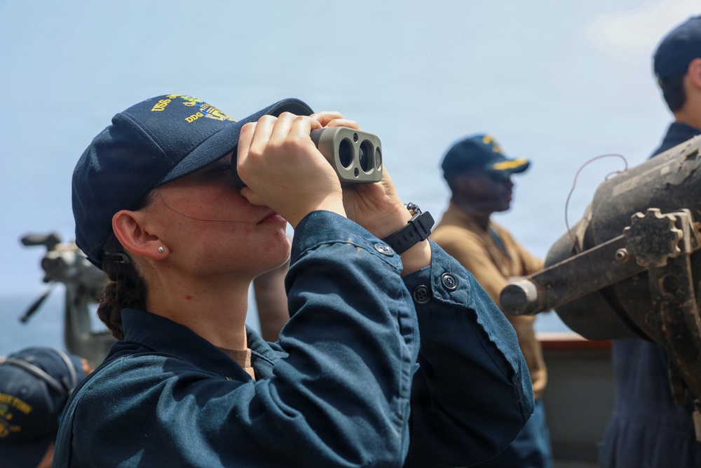 A female sailor aboard a U.S. Navy vessel uses binoculars to scan the horizon while on deck watch. She is wearing a navy blue uniform and a USS ship cap with 'DDG' designation visible. At least two other crew members are visible in the background, also in Navy working uniforms.