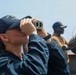 A female sailor aboard a U.S. Navy vessel uses binoculars to scan the horizon while on deck watch. She is wearing a navy blue uniform and a USS ship cap with 'DDG' designation visible. At least two other crew members are visible in the background, also in Navy working uniforms.
