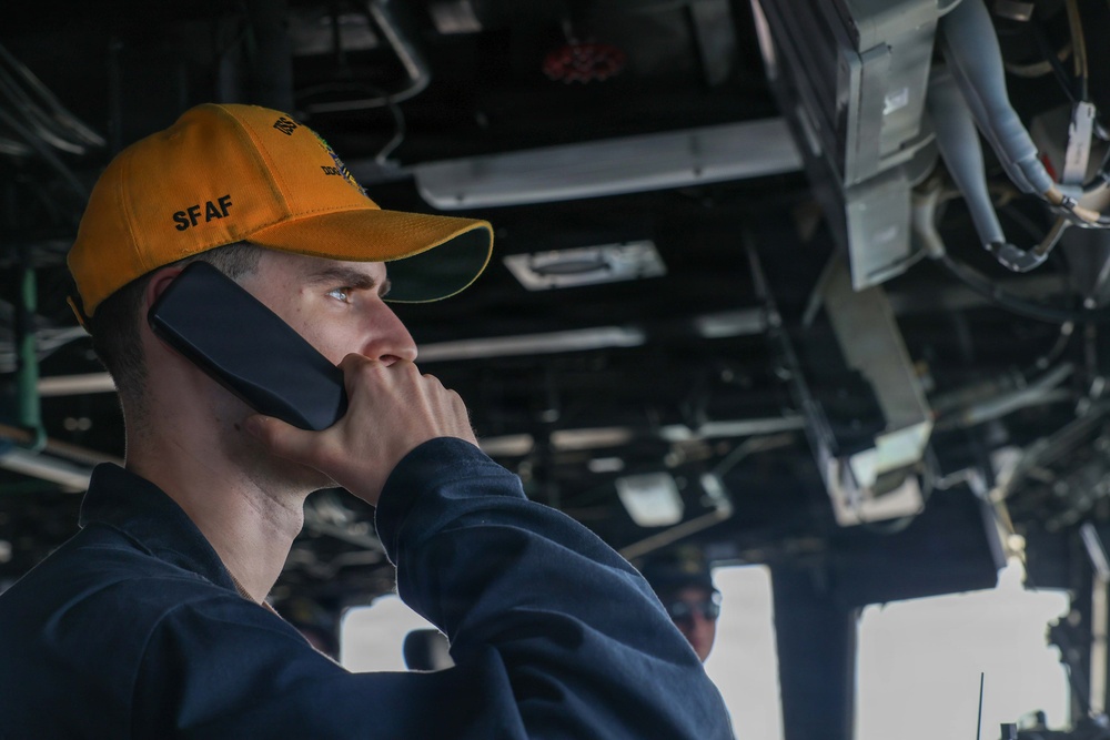 A young male personnel wearing an orange/yellow cap labeled 'SFAF' is holding a communication handset to his ear while looking outward. He appears to be aboard a naval vessel, likely on the bridge or a command station, surrounded by overhead equipment, cables, and machinery. The setting suggests a military maritime environment with operational equipment visible in the background.