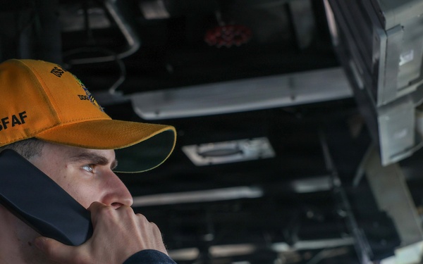 A young male personnel wearing an orange/yellow cap labeled 'SFAF' is holding a communication handset to his ear while looking outward. He appears to be aboard a naval vessel, likely on the bridge or a command station, surrounded by overhead equipment, cables, and machinery. The setting suggests a military maritime environment with operational equipment visible in the background.