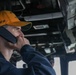 A young male personnel wearing an orange/yellow cap labeled 'SFAF' is holding a communication handset to his ear while looking outward. He appears to be aboard a naval vessel, likely on the bridge or a command station, surrounded by overhead equipment, cables, and machinery. The setting suggests a military maritime environment with operational equipment visible in the background.