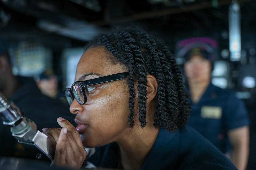 A woman wearing black-framed glasses and a dark uniform is leaning forward and looking intently at an instrument or display, with her hand raised near her chin in a thoughtful pose. The setting appears to be a naval vessel's bridge or operations room, indicated by the equipment and uniformed personnel visible in the background. A second uniformed individual is visible out of focus in the background.