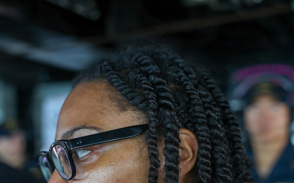 A woman wearing black-framed glasses and a dark uniform is leaning forward and looking intently at an instrument or display, with her hand raised near her chin in a thoughtful pose. The setting appears to be a naval vessel's bridge or operations room, indicated by the equipment and uniformed personnel visible in the background. A second uniformed individual is visible out of focus in the background.