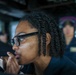A woman wearing black-framed glasses and a dark uniform is leaning forward and looking intently at an instrument or display, with her hand raised near her chin in a thoughtful pose. The setting appears to be a naval vessel's bridge or operations room, indicated by the equipment and uniformed personnel visible in the background. A second uniformed individual is visible out of focus in the background.
