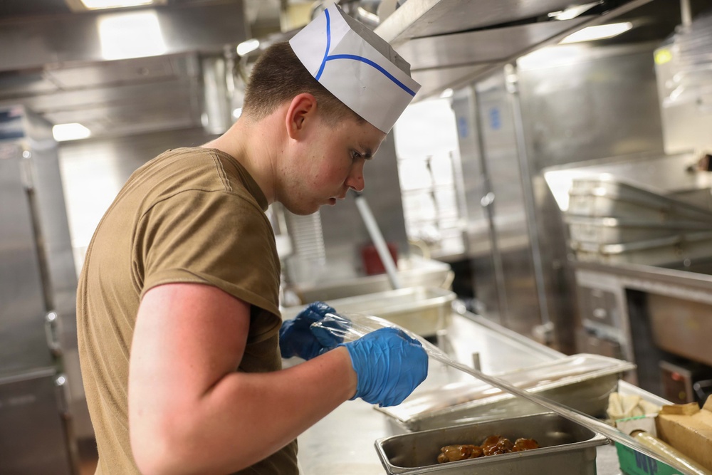 A young person wearing a white paper chef's hat with blue markings and a olive/tan military-style t-shirt is working in a commercial kitchen. They are wearing blue nitrile food-handling gloves and appear to be plating or serving food from a stainless steel hotel pan. The setting appears to be a military mess hall or institutional kitchen based on the equipment and attire.