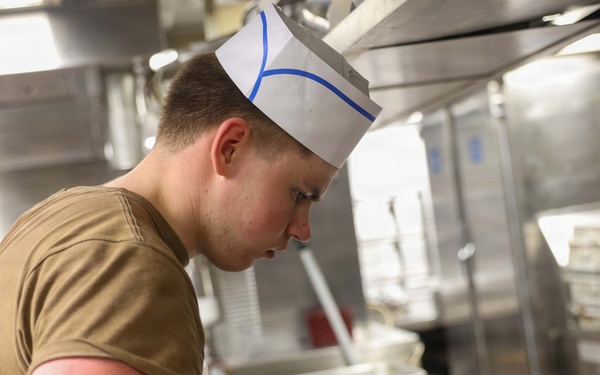 A young person wearing a white paper chef's hat with blue markings and a olive/tan military-style t-shirt is working in a commercial kitchen. They are wearing blue nitrile food-handling gloves and appear to be plating or serving food from a stainless steel hotel pan. The setting appears to be a military mess hall or institutional kitchen based on the equipment and attire.