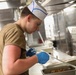 A young person wearing a white paper chef's hat with blue markings and a olive/tan military-style t-shirt is working in a commercial kitchen. They are wearing blue nitrile food-handling gloves and appear to be plating or serving food from a stainless steel hotel pan. The setting appears to be a military mess hall or institutional kitchen based on the equipment and attire.
